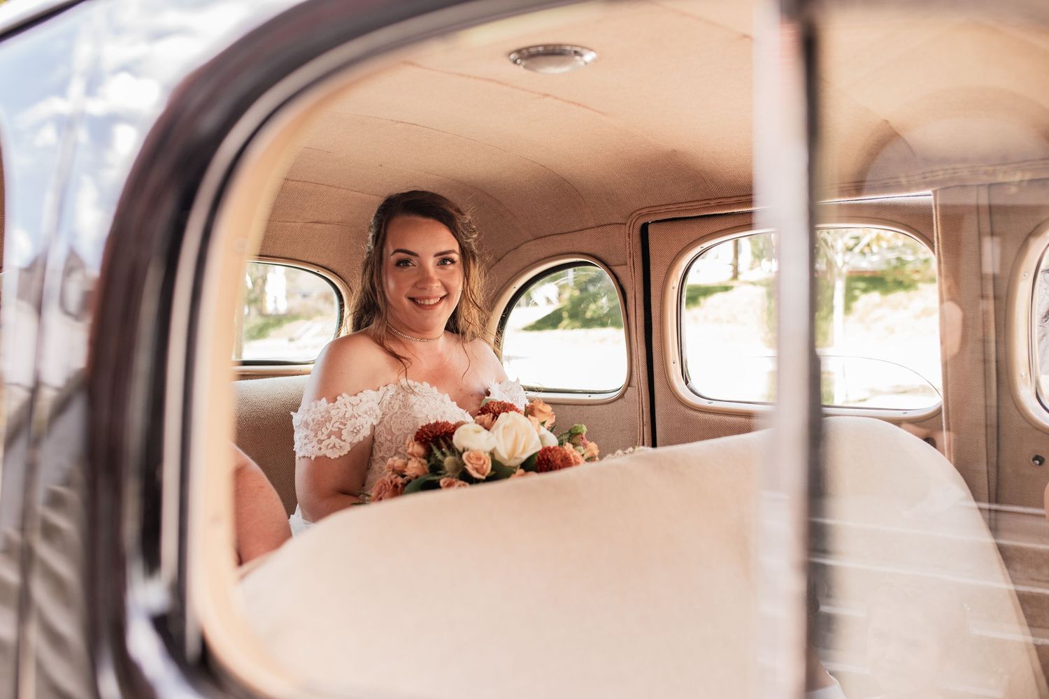 View through car window of bride in vintage automobile holding bouquet.