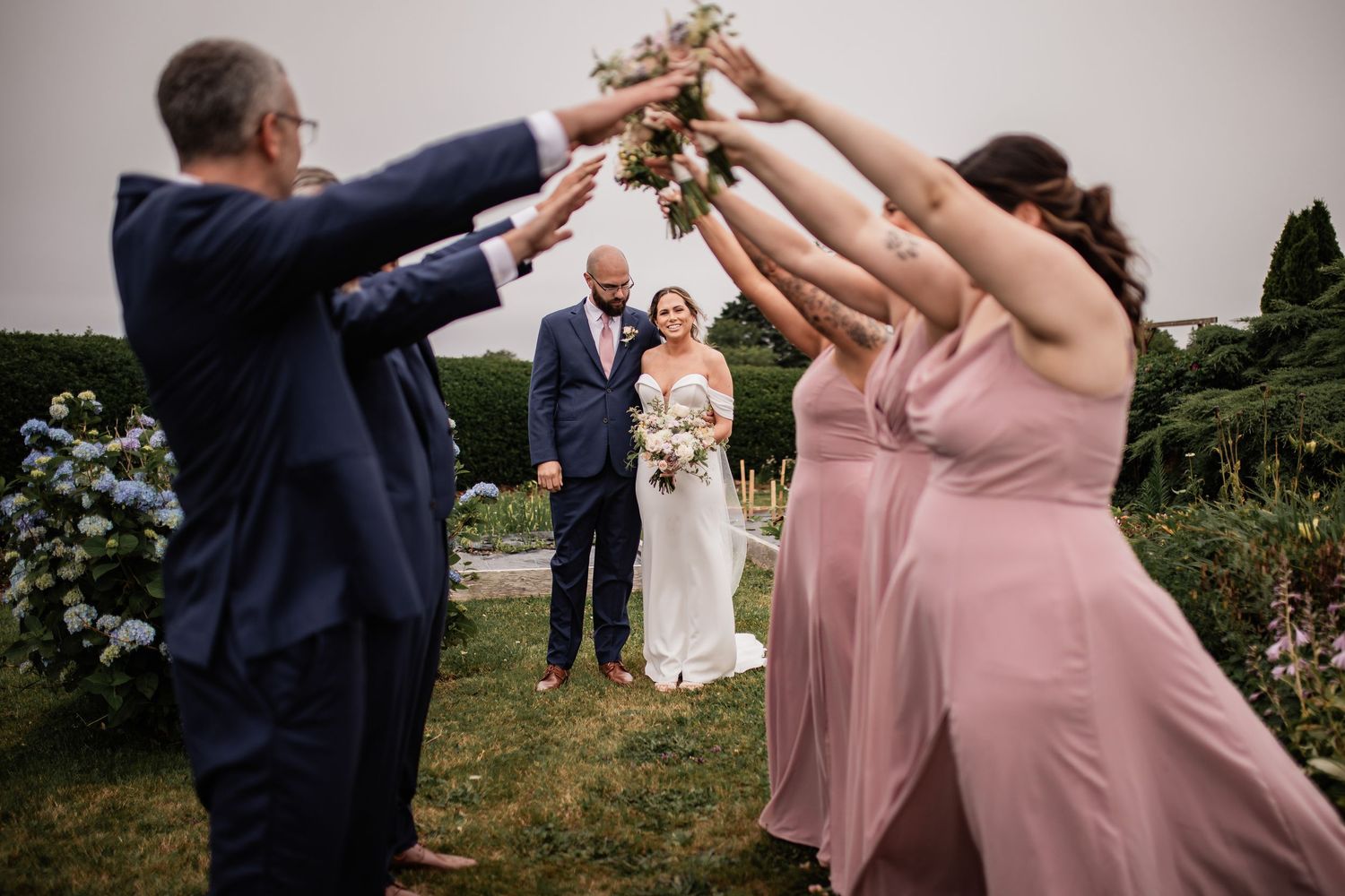 Wedding party creates tunnel of raised arms while couple stands in background.