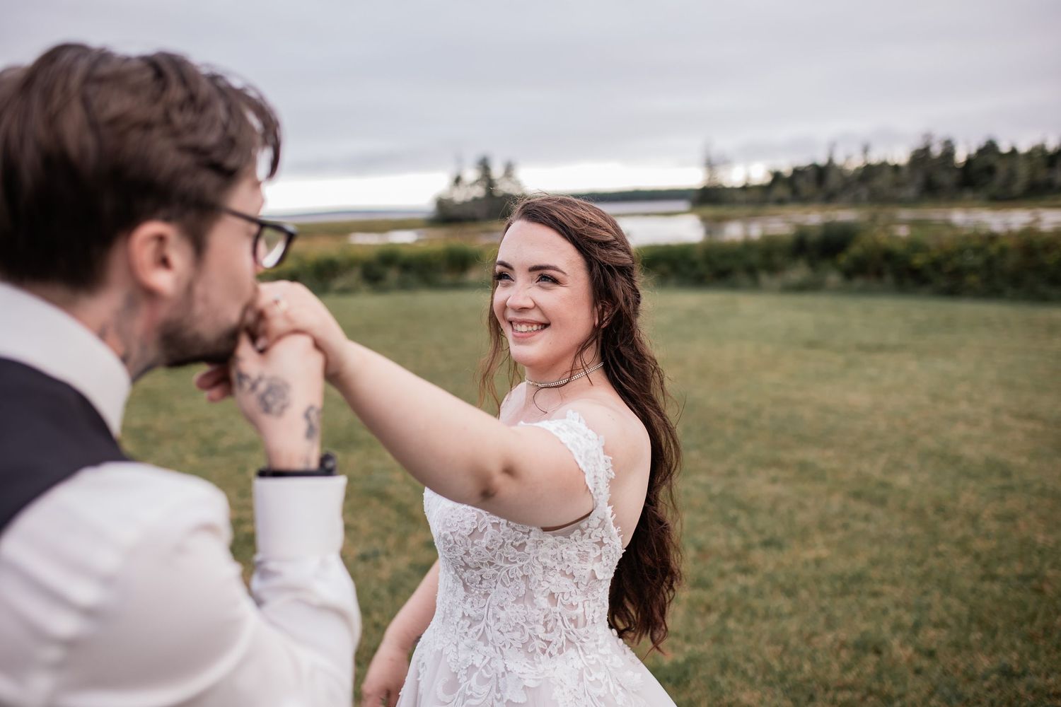 Romantic outdoor wedding moment with groom kissing bride's hand in grassy field.