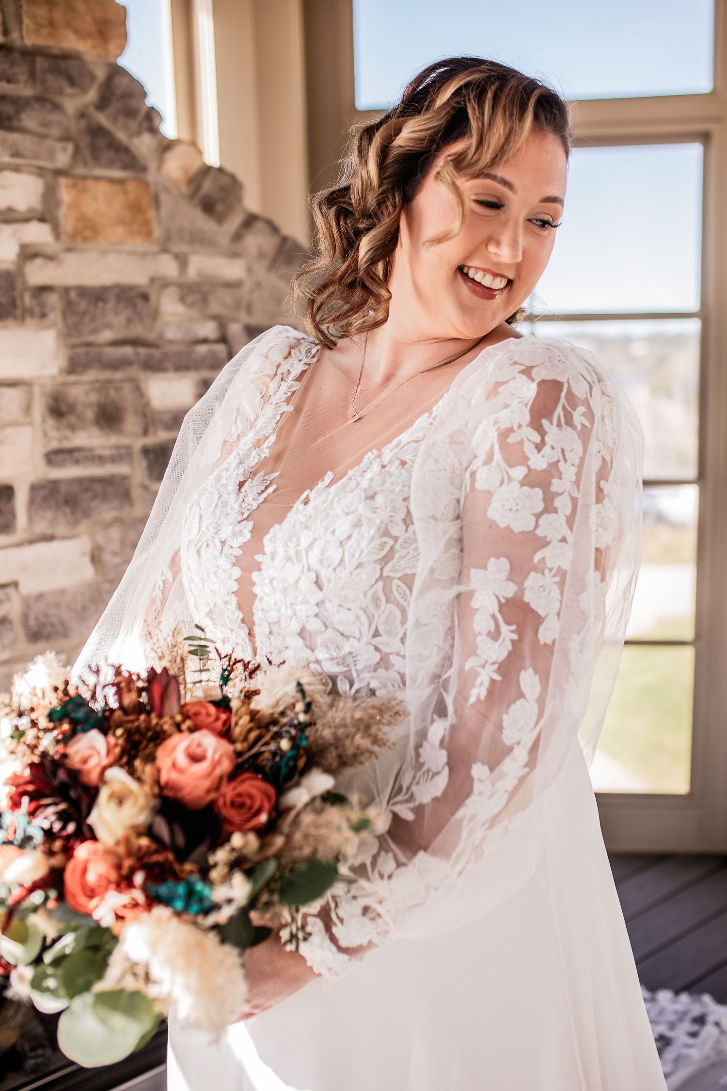 Bride wearing lace long sleeve wedding dress holding coral and white bouquet while smiling.