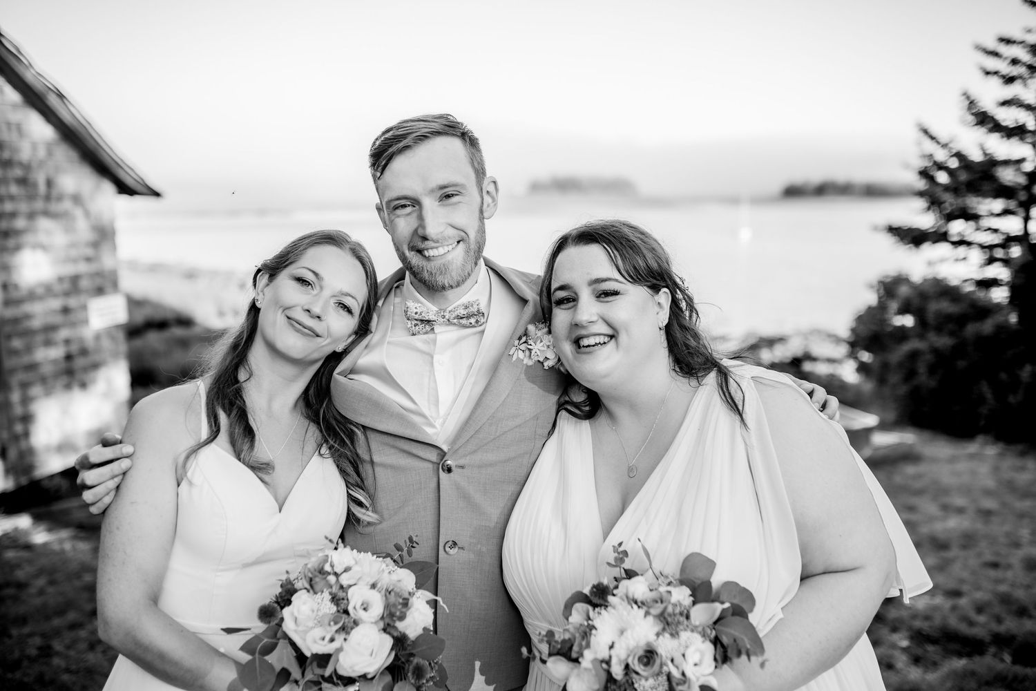 A black and white wedding photo showing three people embracing outdoors while holding flower bouquets.