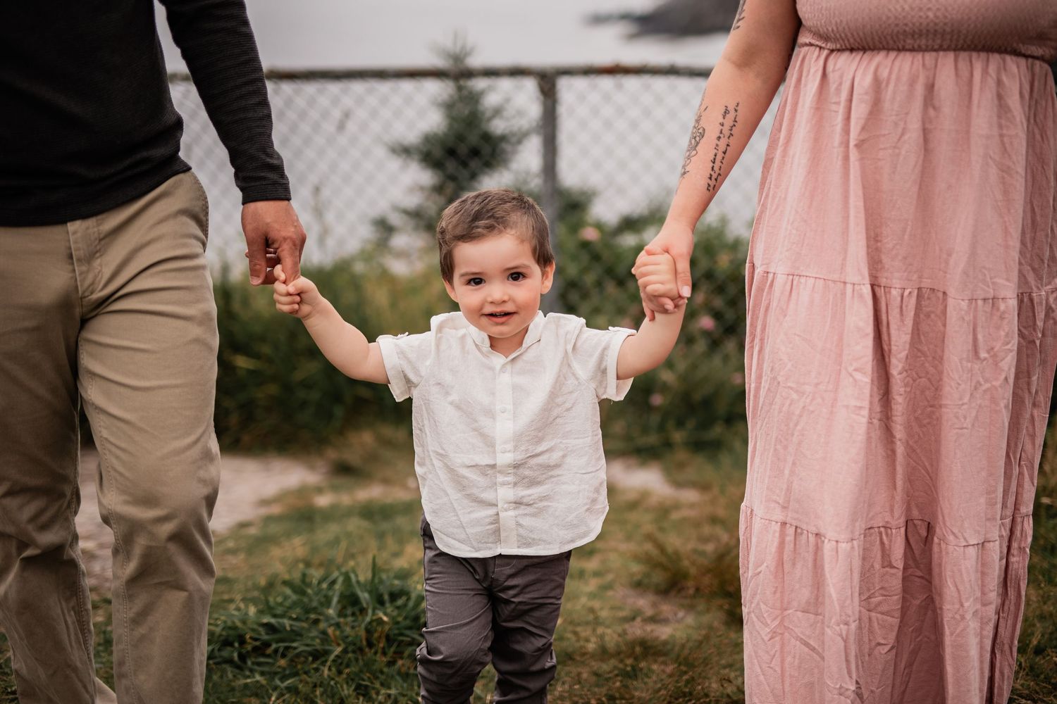 Young child in white shirt walking while holding parents' hands on grassy path.