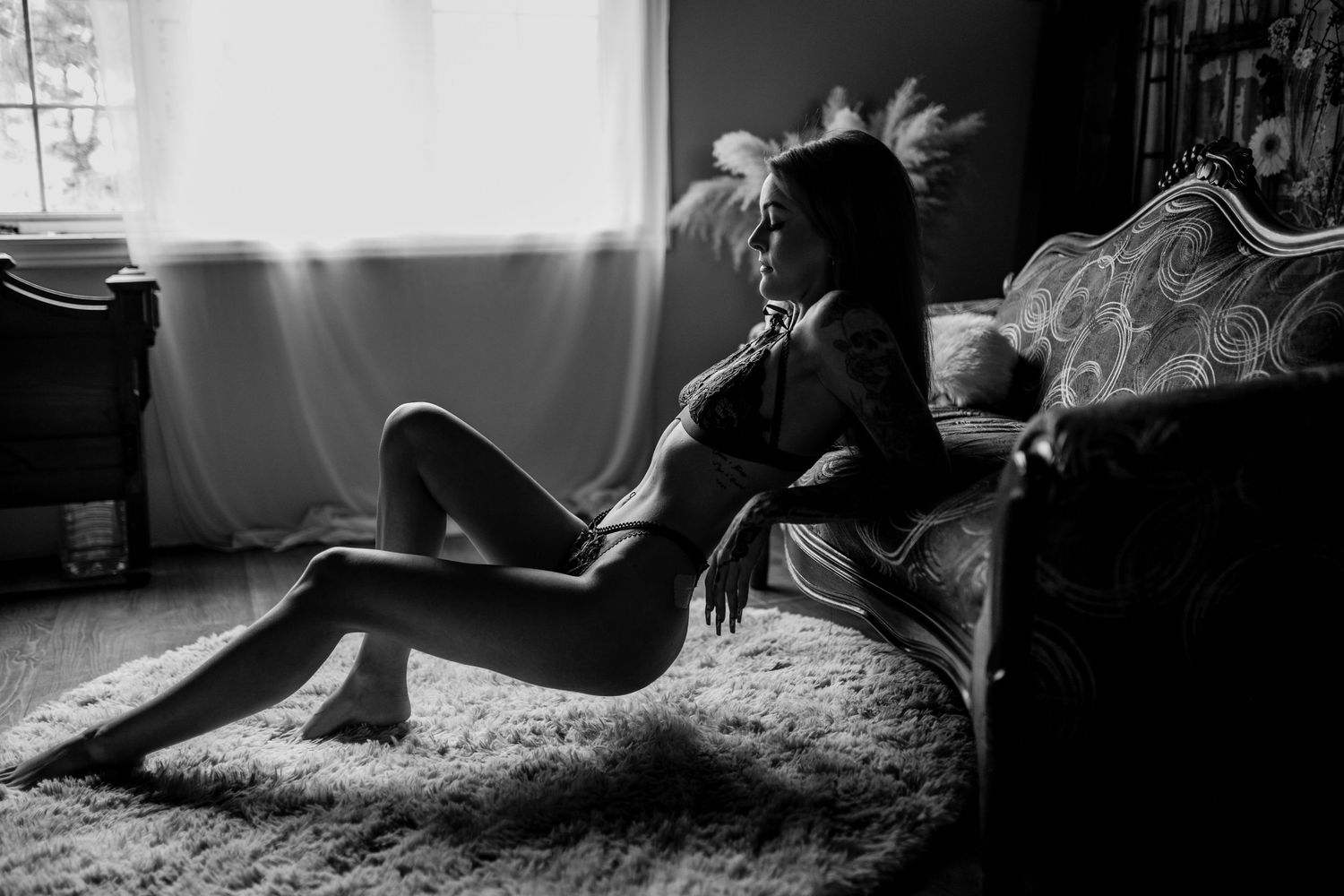 A black and white artistic portrait of someone sitting on the floor beside a bed while sunlight streams through a window.