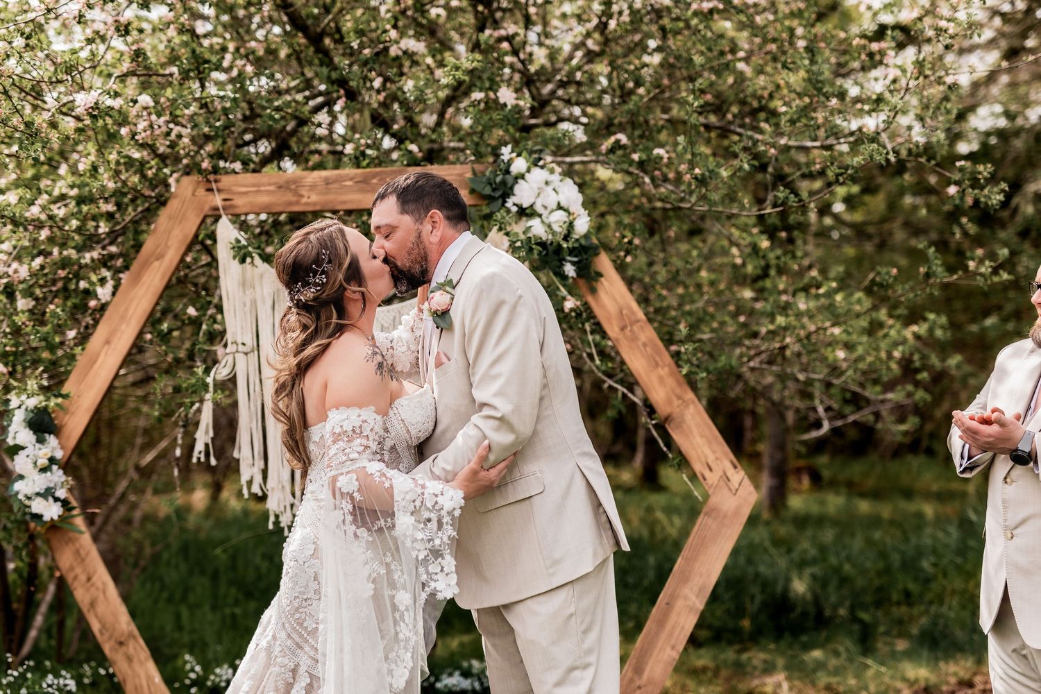 A wedding ceremony under a wooden hexagonal arch with blooming trees in the background.