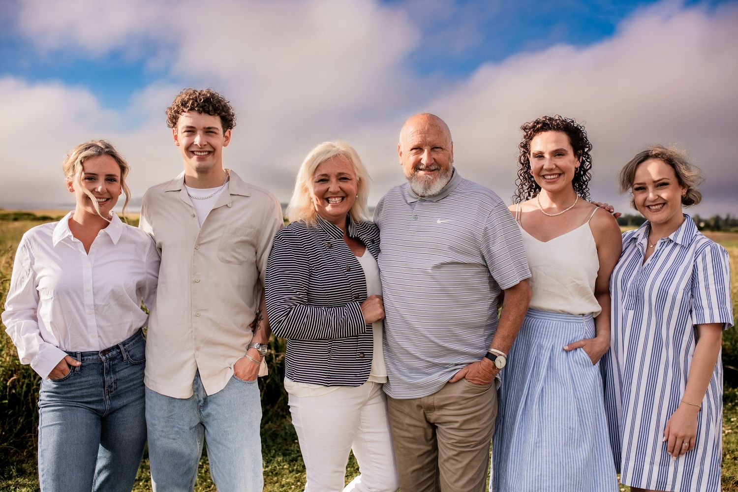 Multi-generational family poses together outdoors on a sunny day against cloudy sky.
