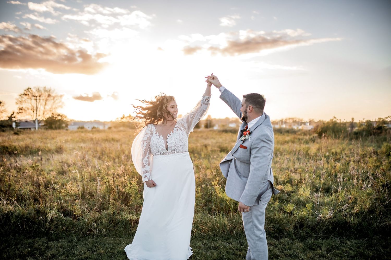 A couple dressed in wedding attire dance joyfully in a sunlit meadow at sunset.