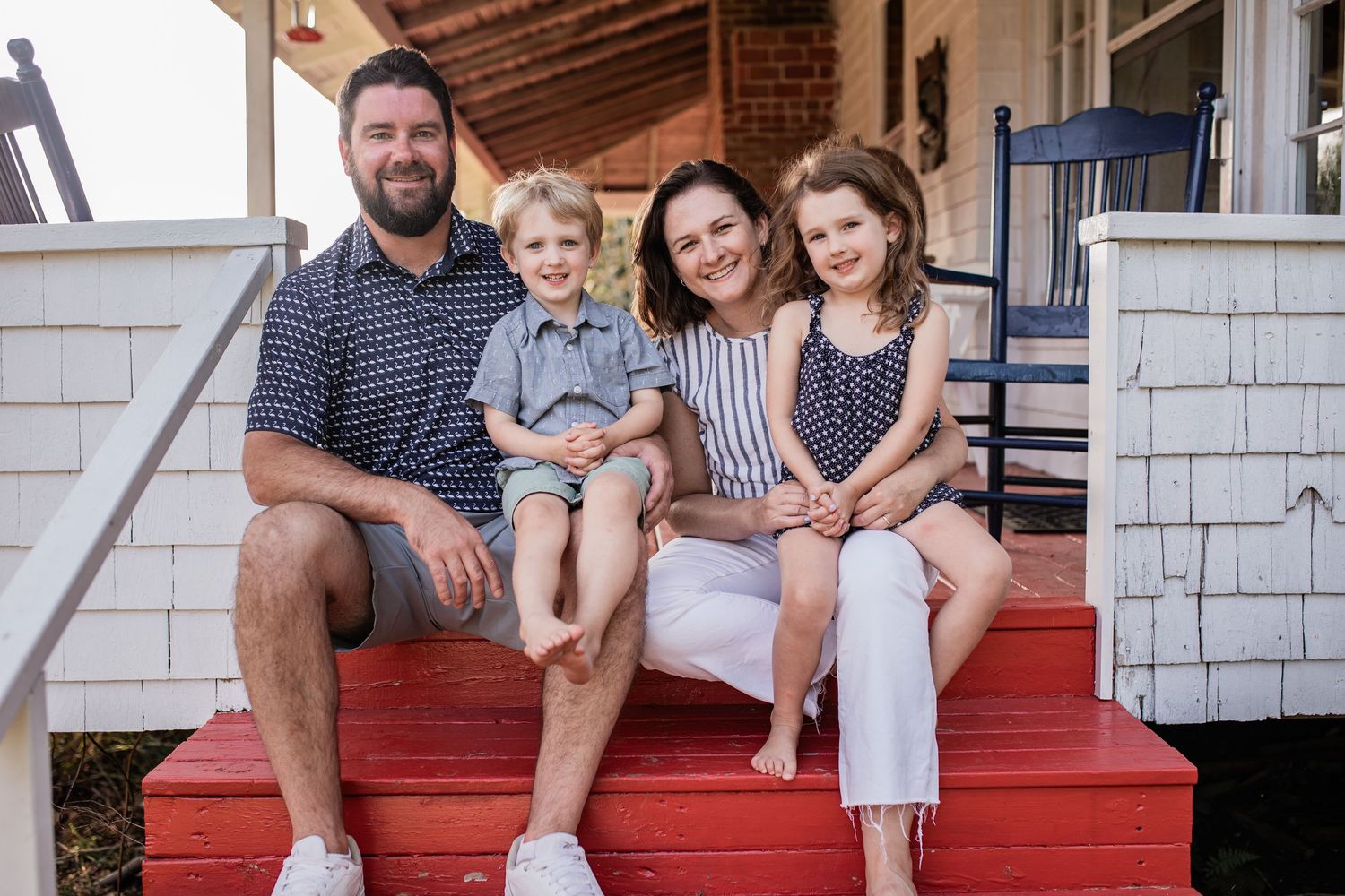 Family of four sitting together on red painted porch steps of a white house.