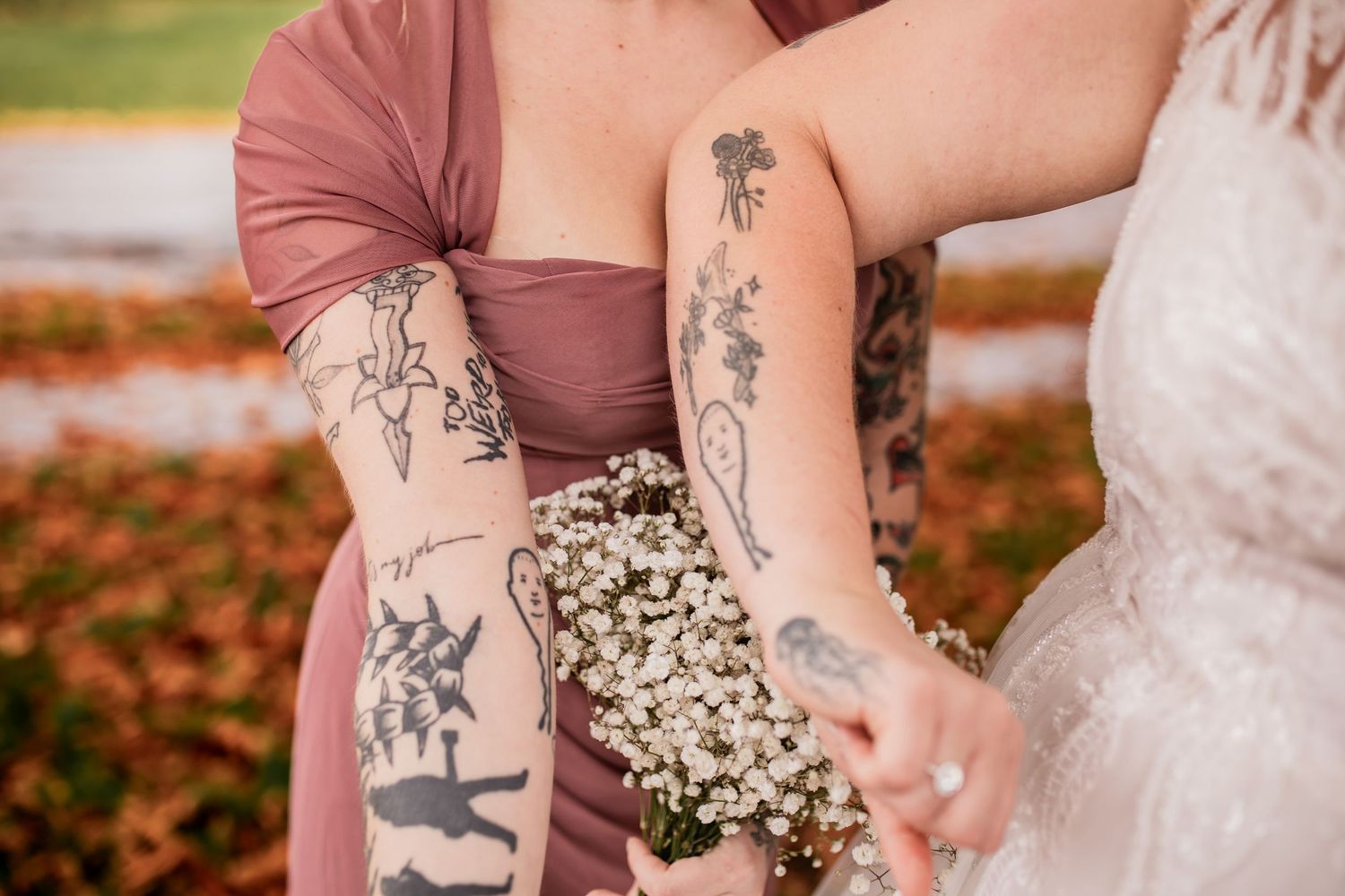 Detailed floral tattoos adorn arms holding delicate baby's breath bouquets at an outdoor wedding.