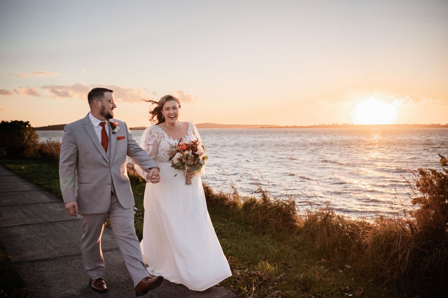 Newlyweds walking along coastal path during sunset with bride holding autumn bouquet.