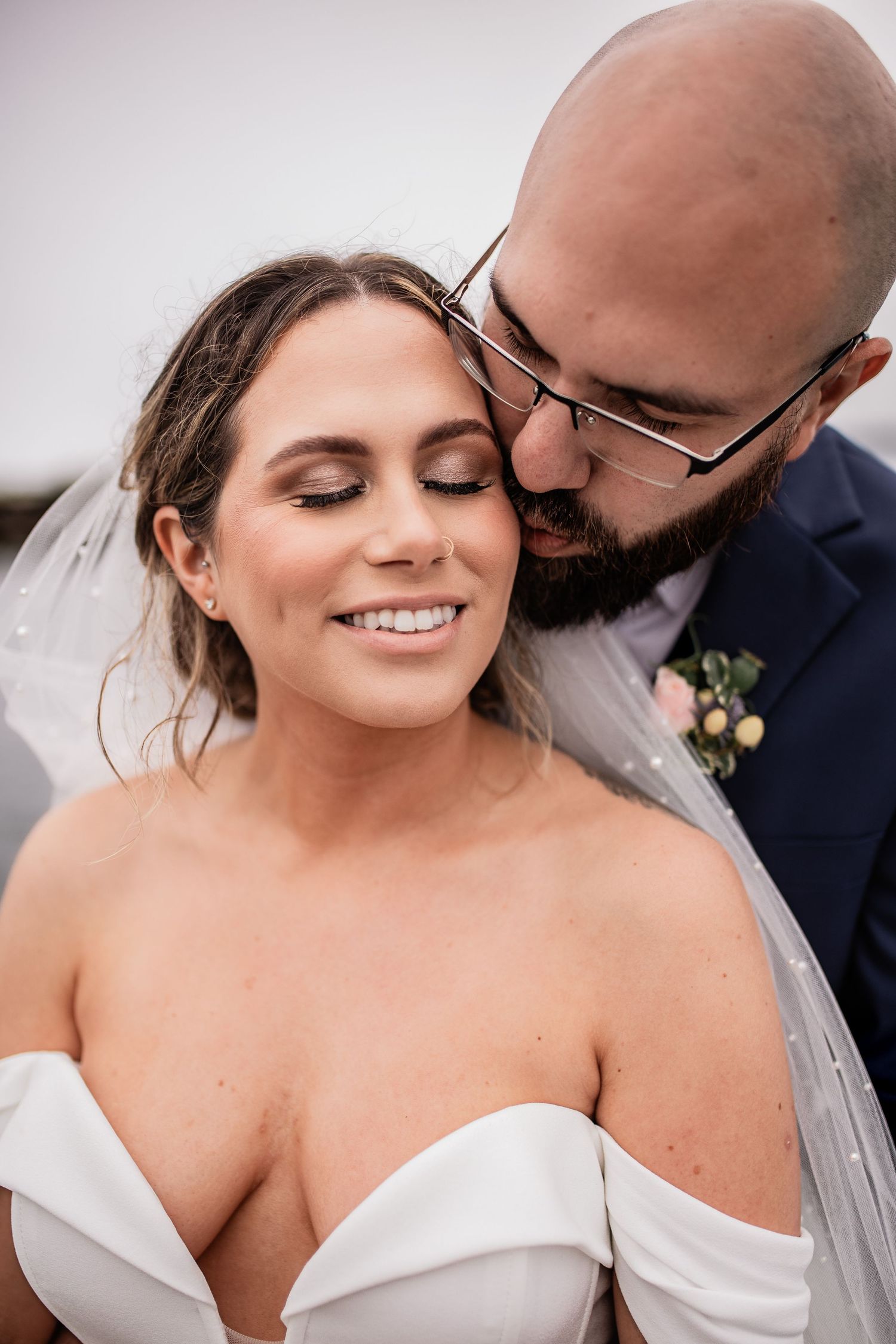 Intimate wedding portrait of couple sharing tender moment while bride wears off-shoulder white gown and groom wears glasses.