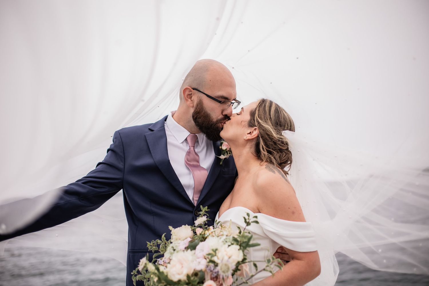 Intimate wedding portrait with flowing veil against cloudy sky.