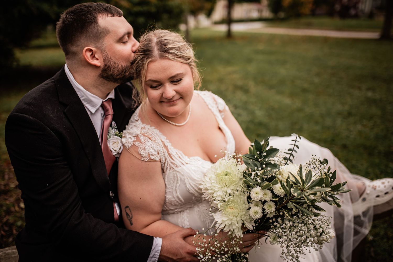 A romantic outdoor wedding moment featuring a couple with a white bouquet on a grassy lawn.