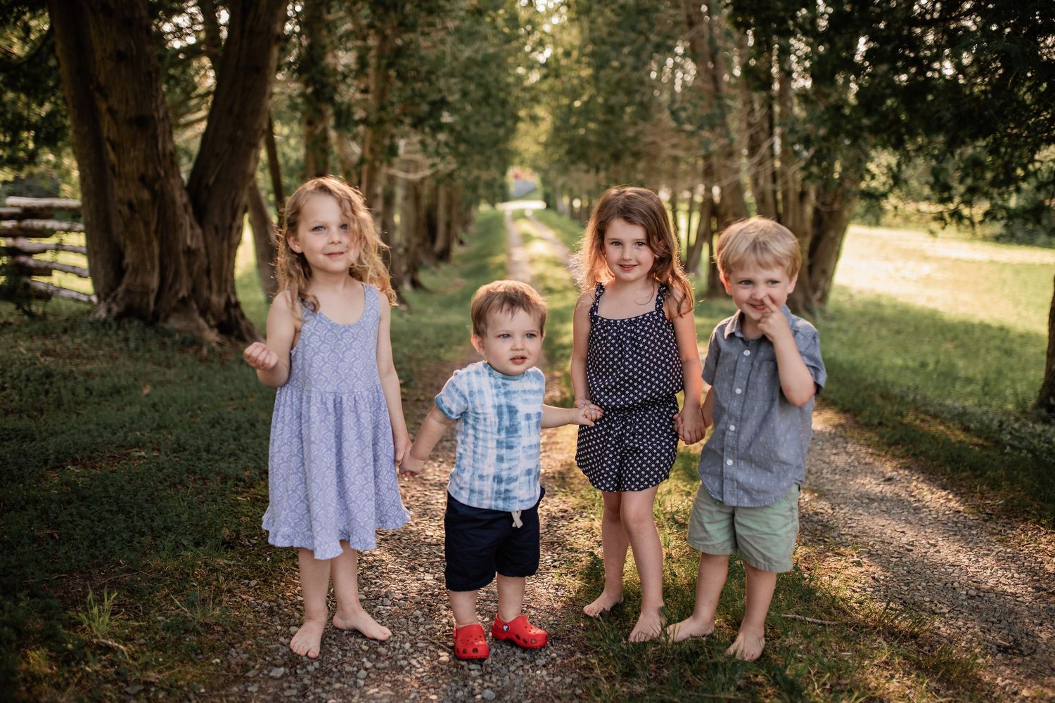 Four young children walking together on a tree-lined dirt path in summer clothing.