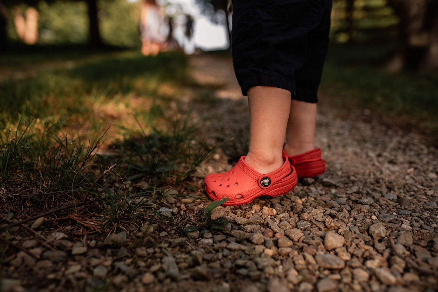 Close-up of feet wearing bright red Crocs sandals walking on a gravel path in a park.