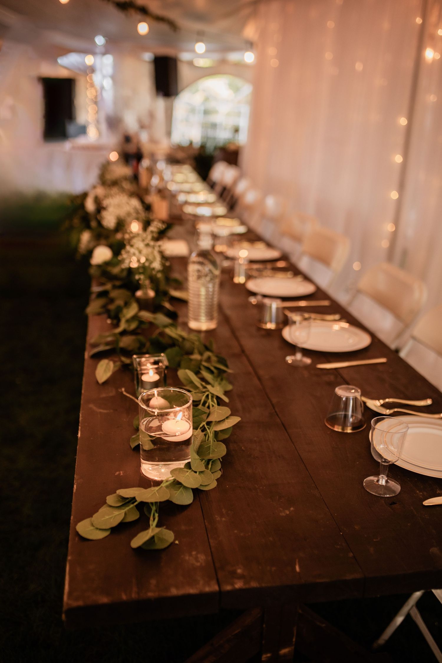 Rustic wooden reception table decorated with greenery, candles and elegant place settings for a wedding.