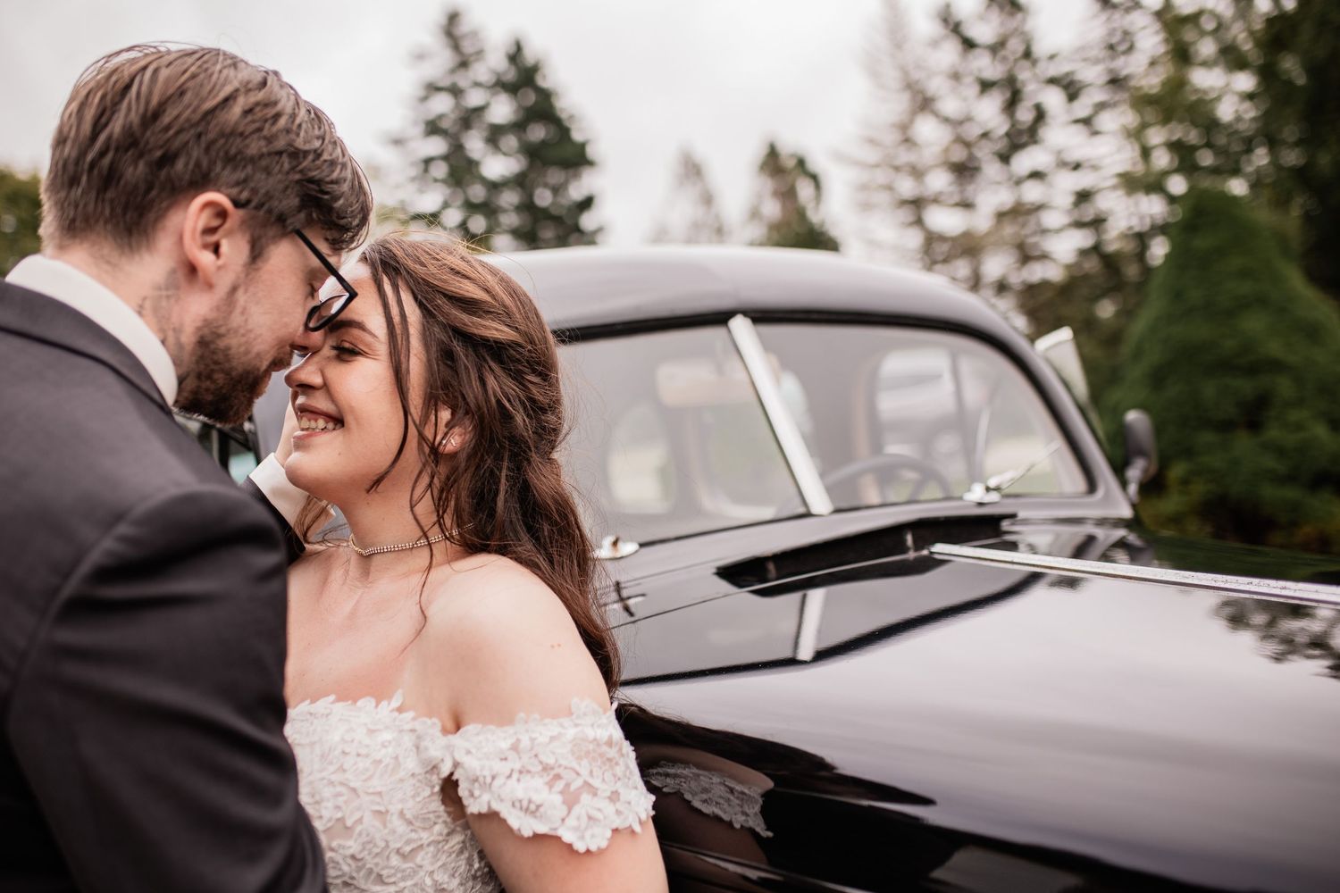 Newlyweds share intimate moment near vintage black car surrounded by evergreen trees.