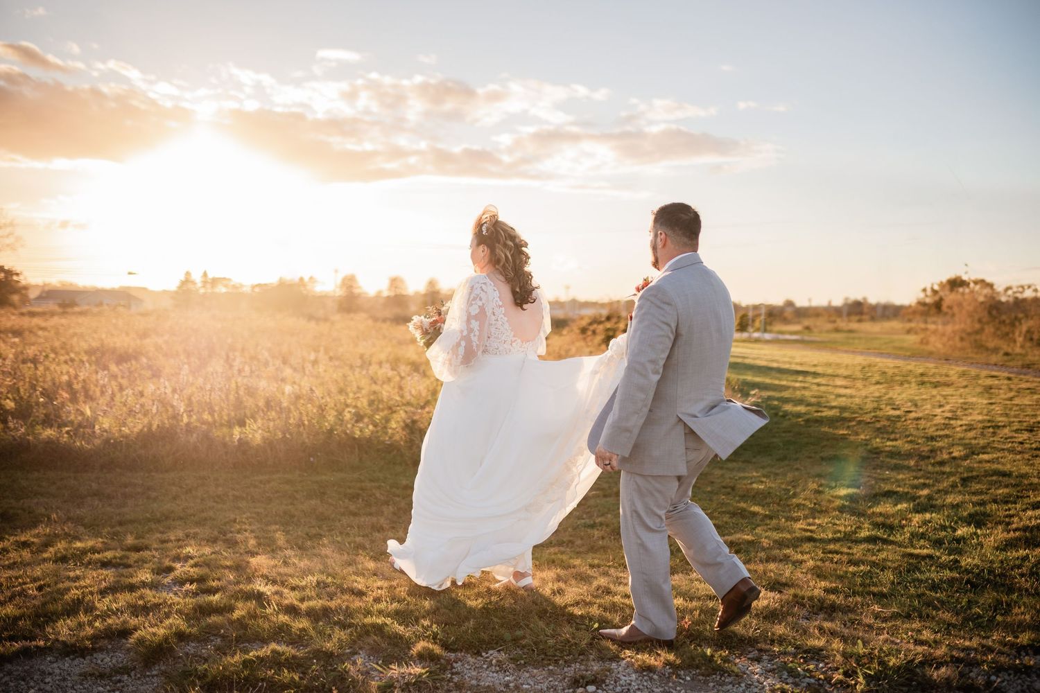 Newlyweds walking hand in hand during golden hour sunset in open field.
