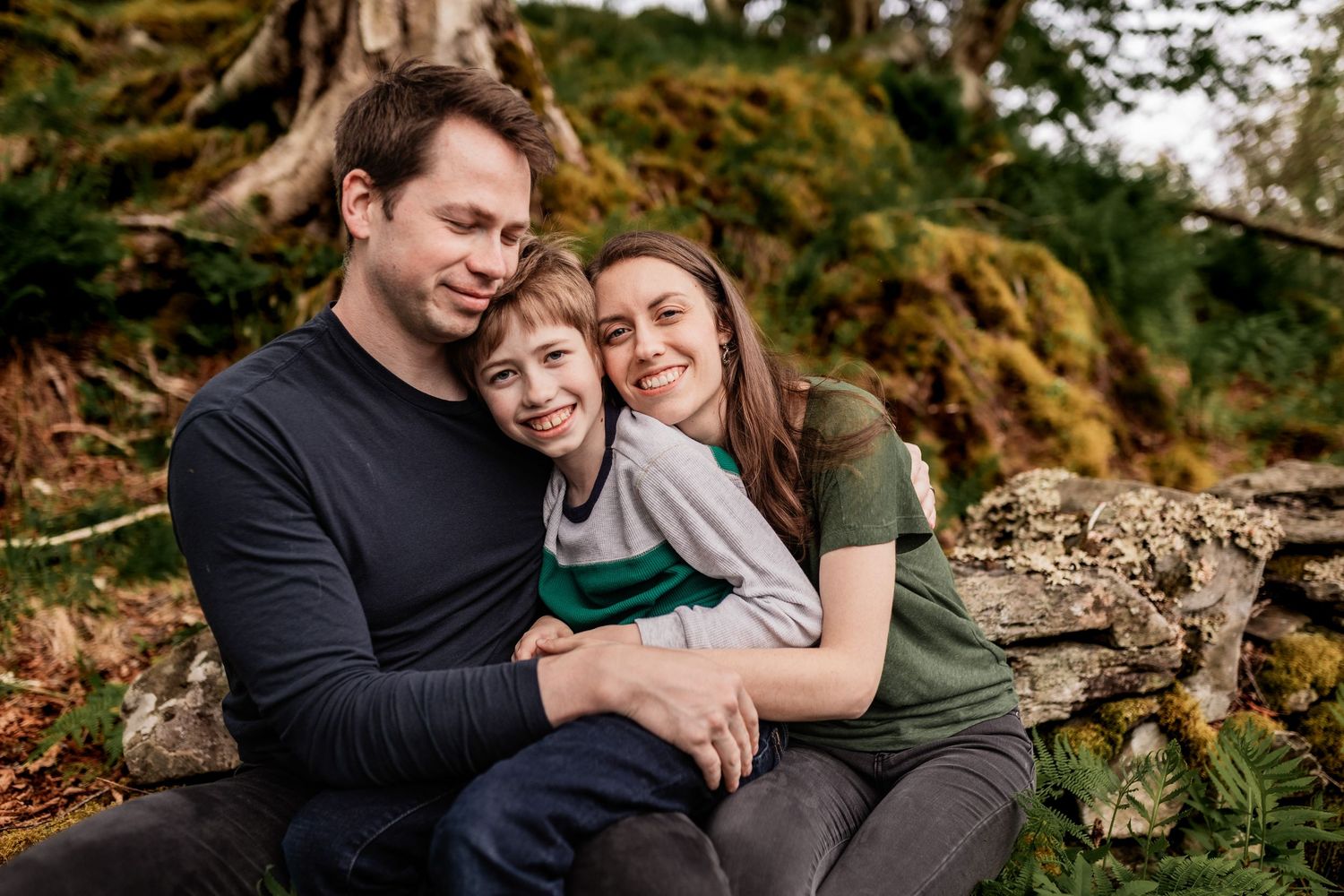Family shares intimate moments while sitting on mossy rocks in a lush green forest setting.