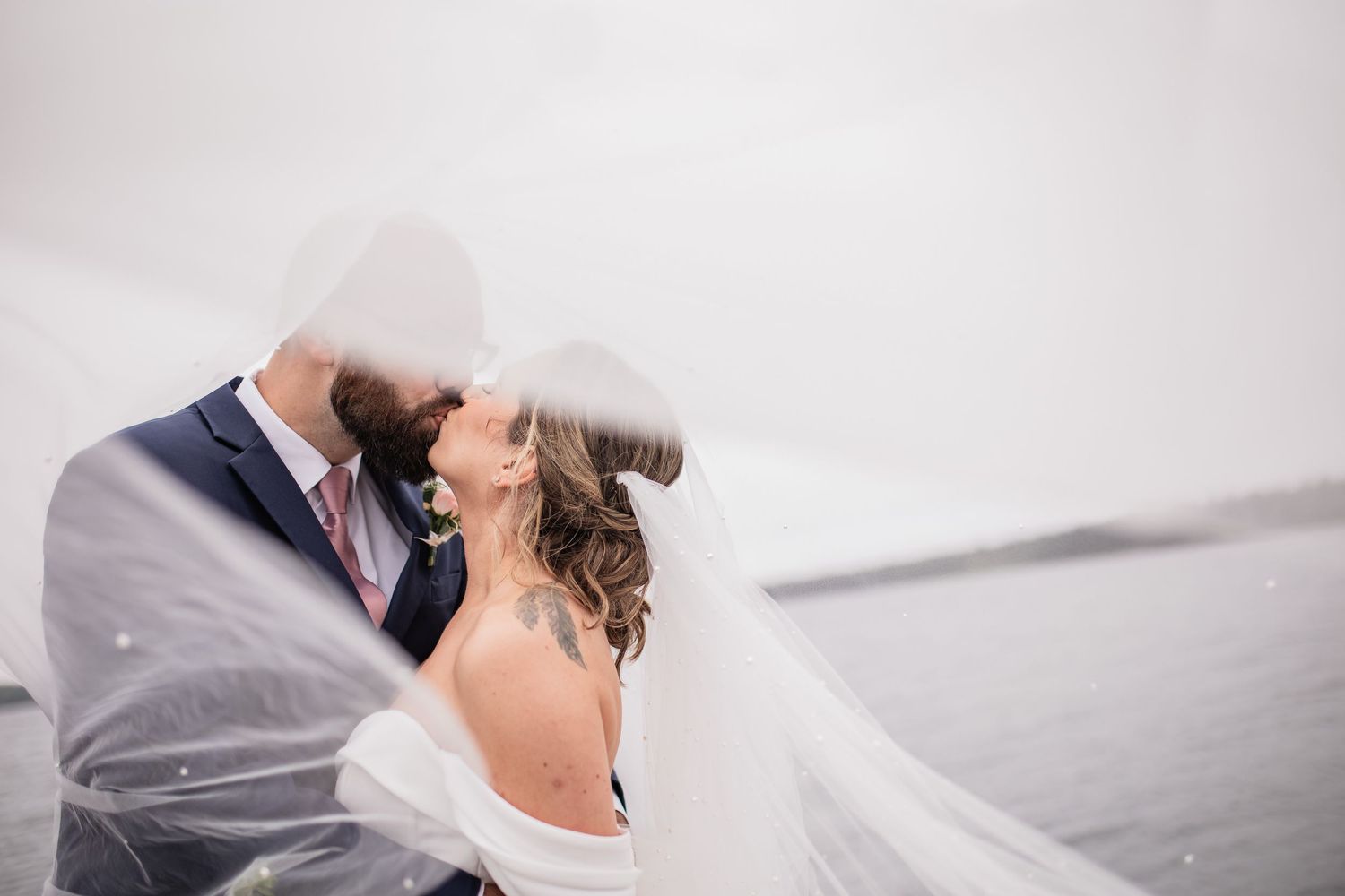 Romantic wedding couple embraces as veil flows in the wind by waterfront.