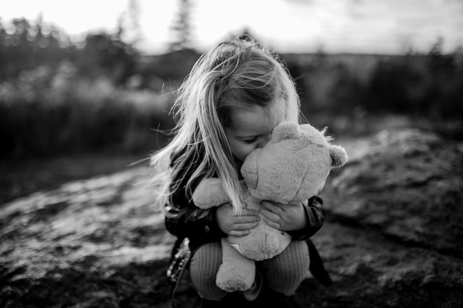 A black and white image of a young one holding a stuffed animal while sitting on rocky terrain.