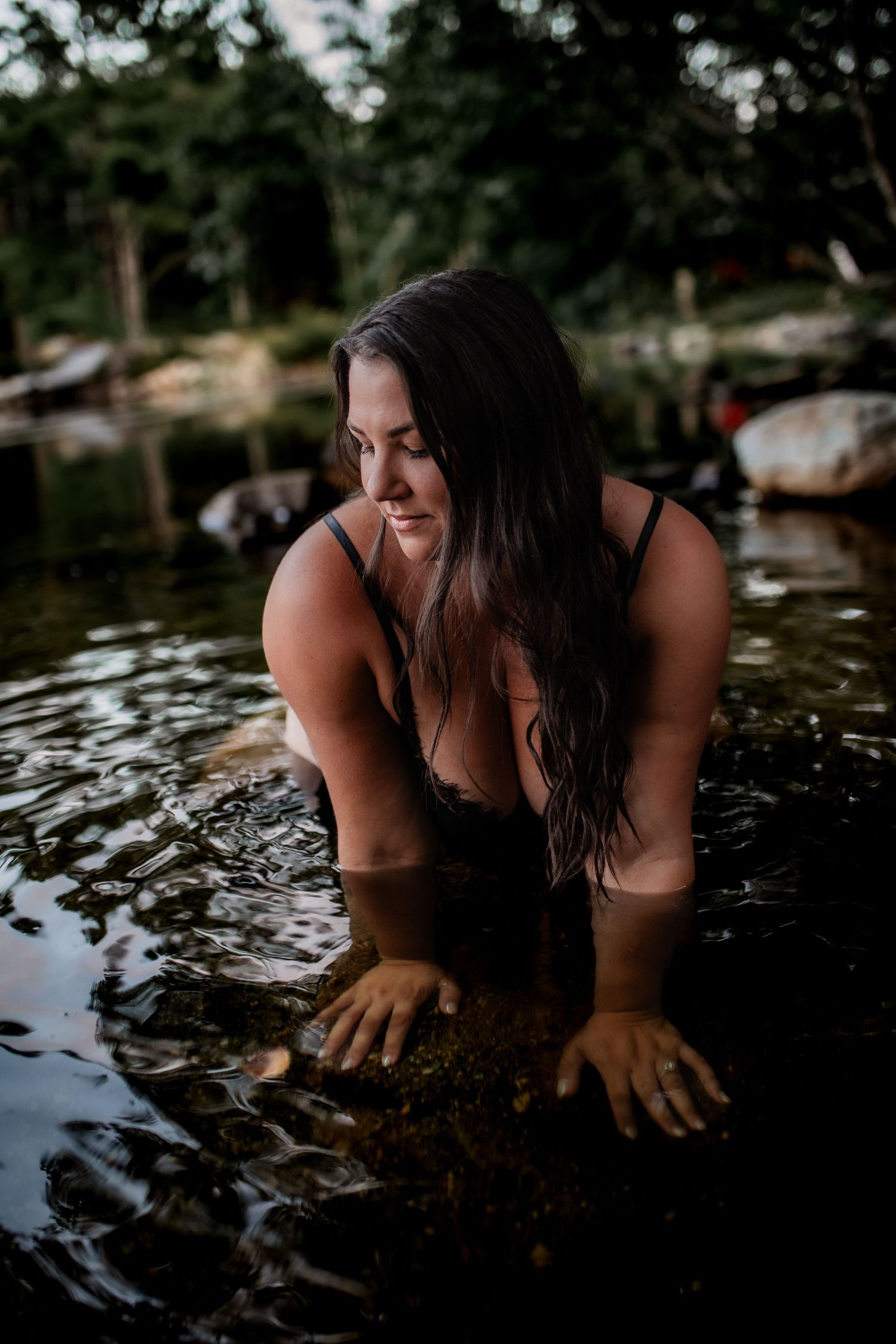 Woman in dark swimwear wading in reflective water at dusk with trees in background.
