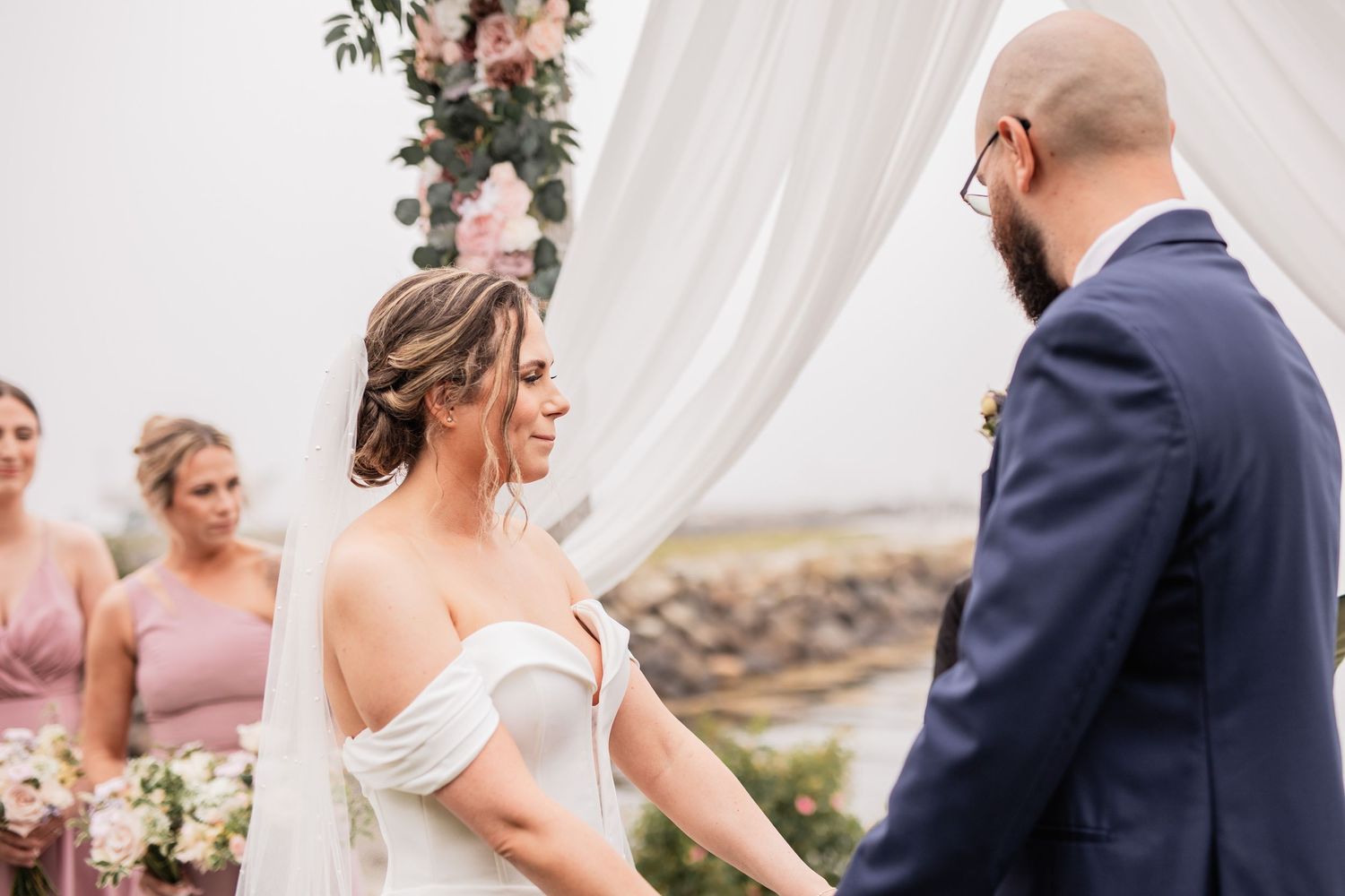Outdoor wedding ceremony with couple exchanging vows under white draping and floral arch.