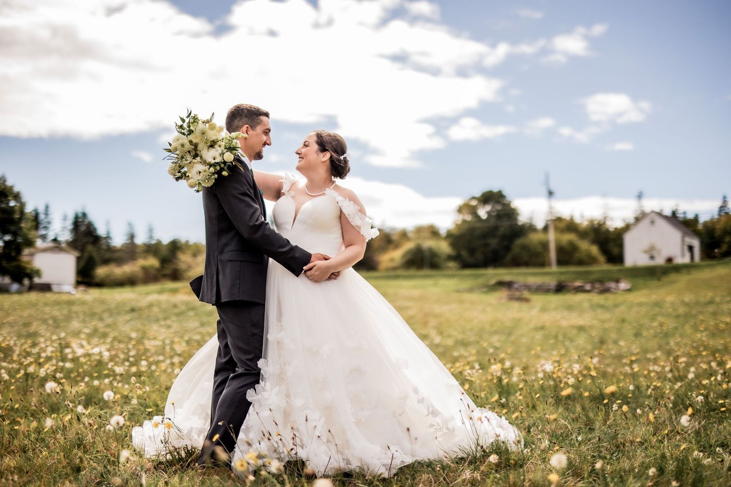 Newlyweds embrace in a sunny wildflower field near a countryside house.