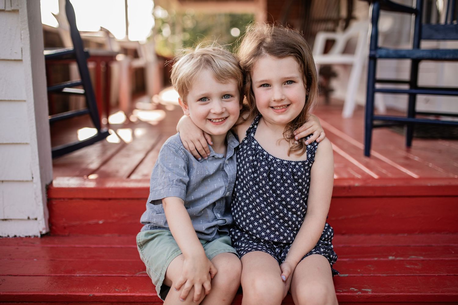 Two young children sitting together on red wooden porch steps on a sunny day.