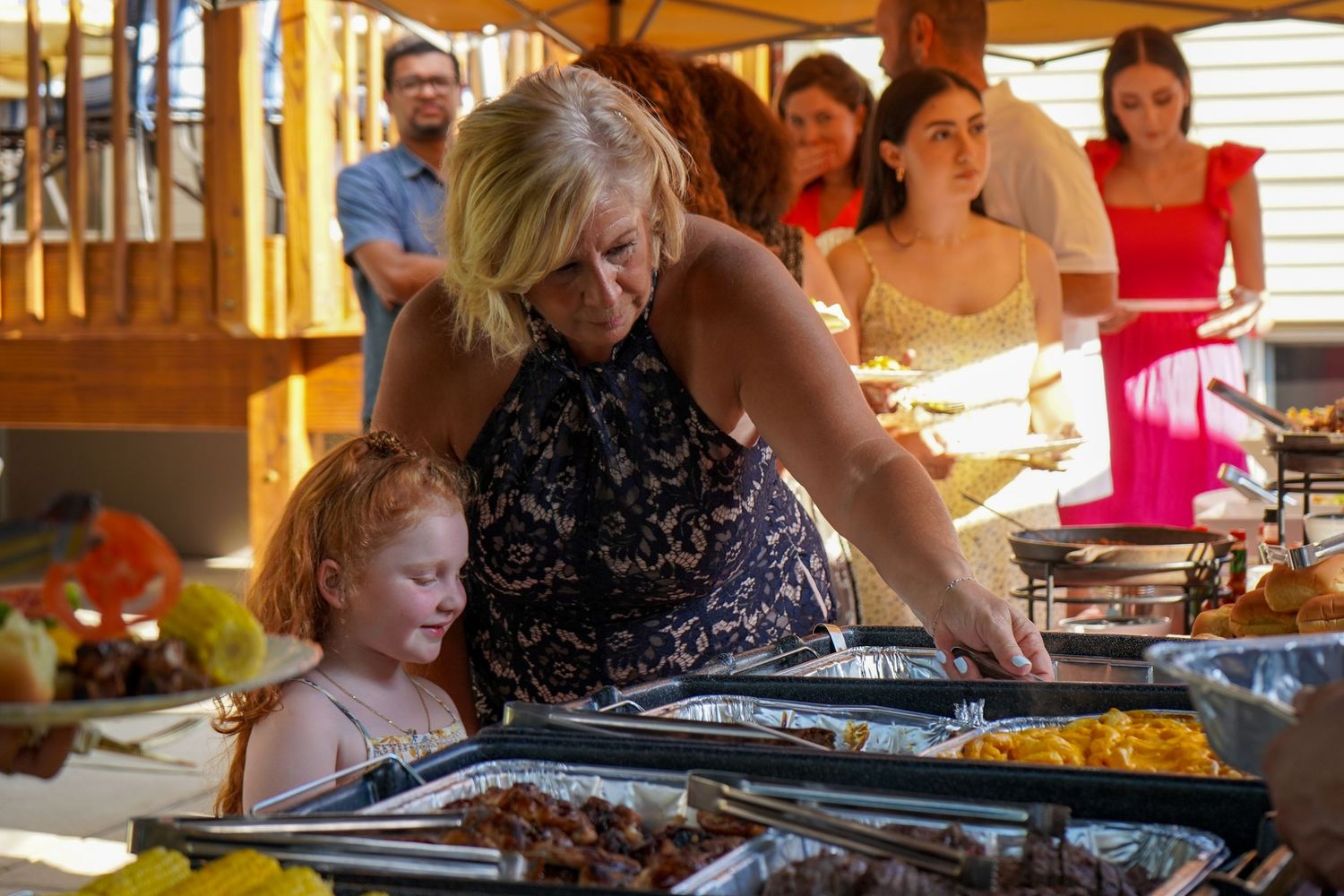 People gather around a buffet serving station with warming trays filled with food at an outdoor event.