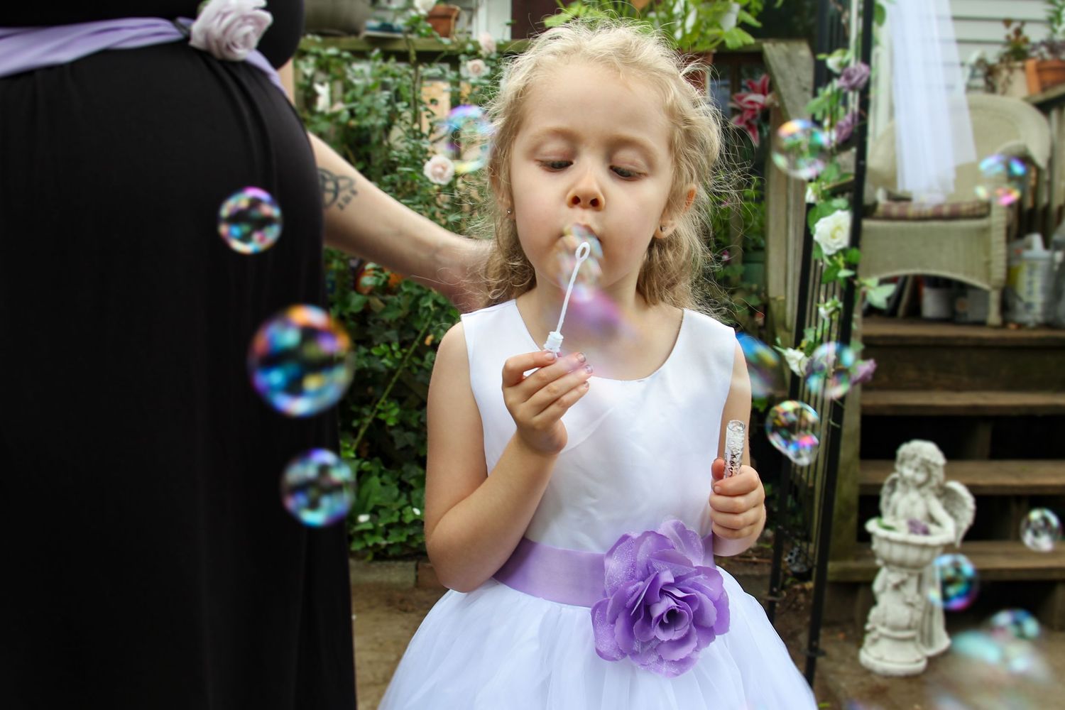 A young child in a white dress blowing bubbles in a garden setting.