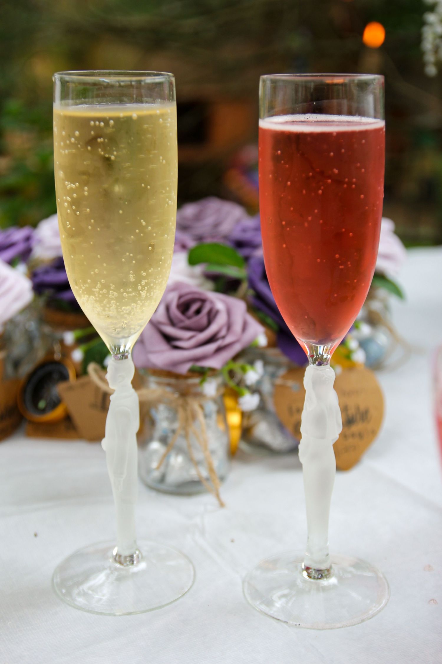 Two champagne flutes filled with sparkling wine next to purple roses on a white tablecloth.