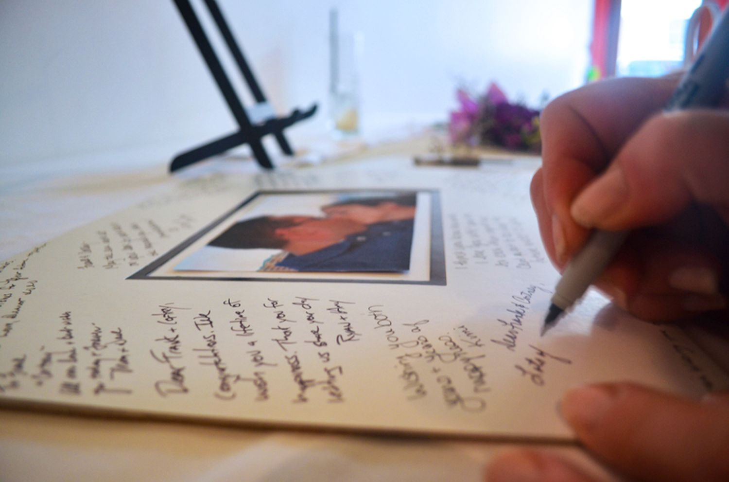 A hand writes in a journal or guest book next to a photograph and decorative elements.