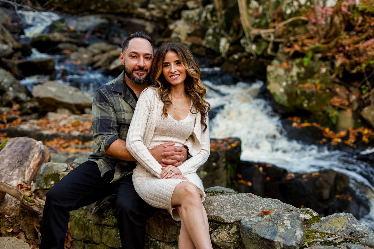 Couple sitting on rocks by rushing stream in autumn forest setting.