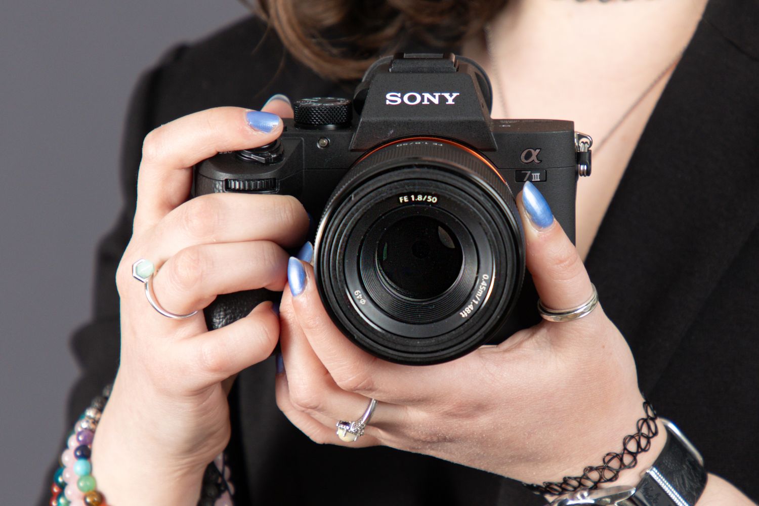 Hands holding Sony Alpha camera with beaded bracelets and blue nail polish detail.