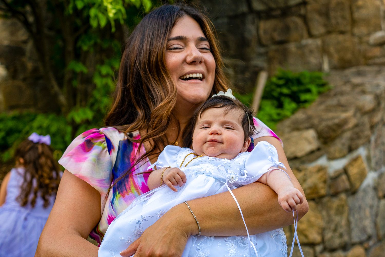 A joyful moment captured between a parent holding a smiling baby dressed in white against a stone wall backdrop.