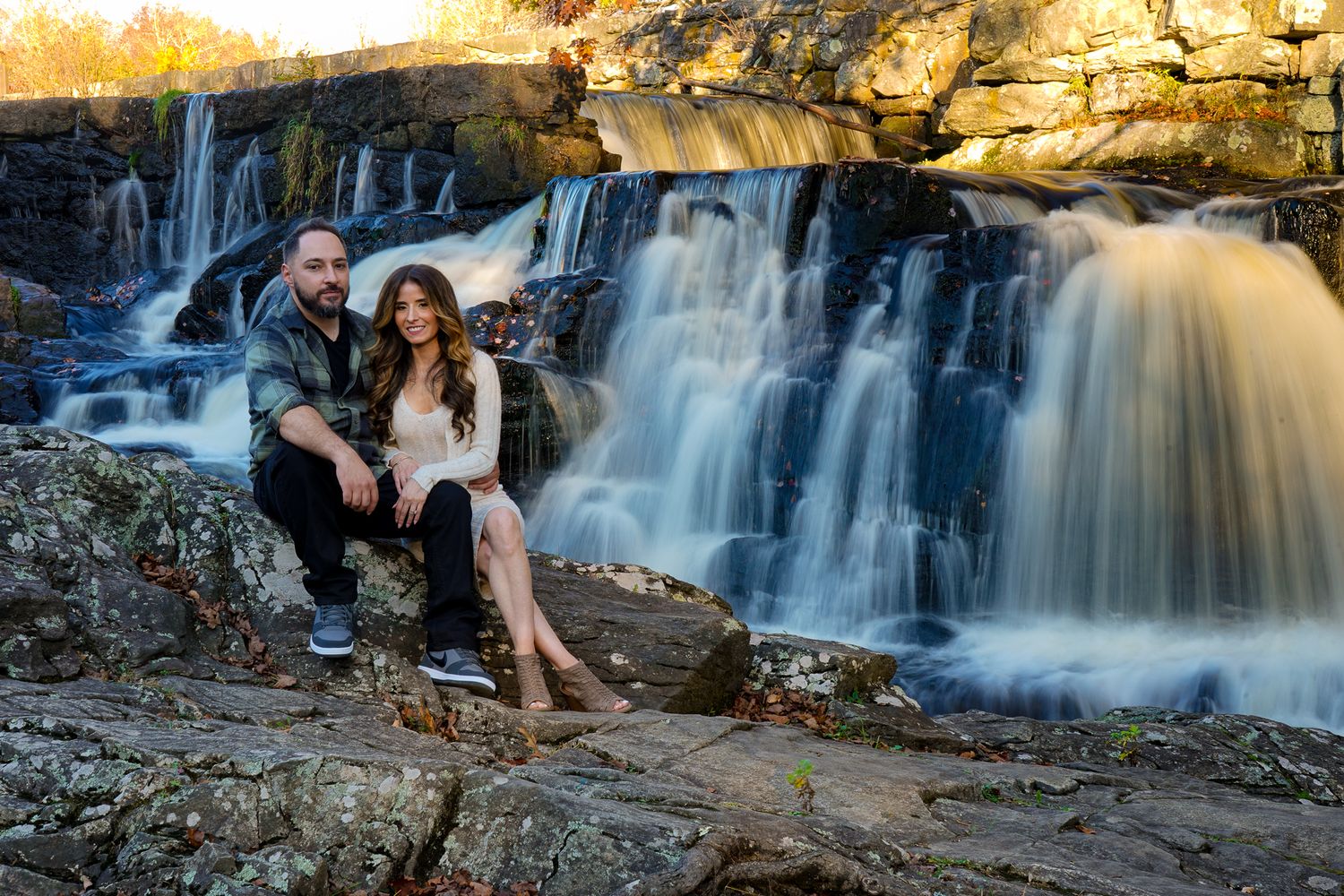 Couple sitting together by cascading waterfall during golden sunset in autumn.