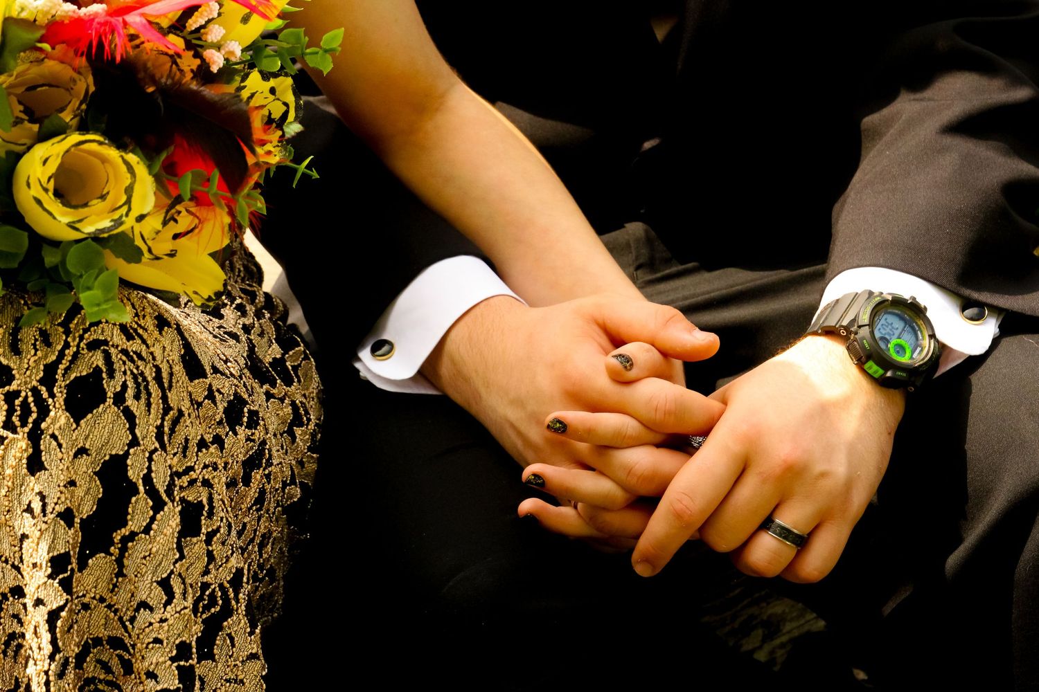 Close-up of joined hands with wedding rings and a wrist watch against formal attire.