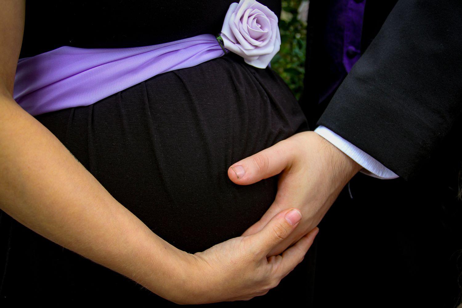 Close up of hands holding a pregnant belly adorned with a purple sash and rose at a formal occasion.