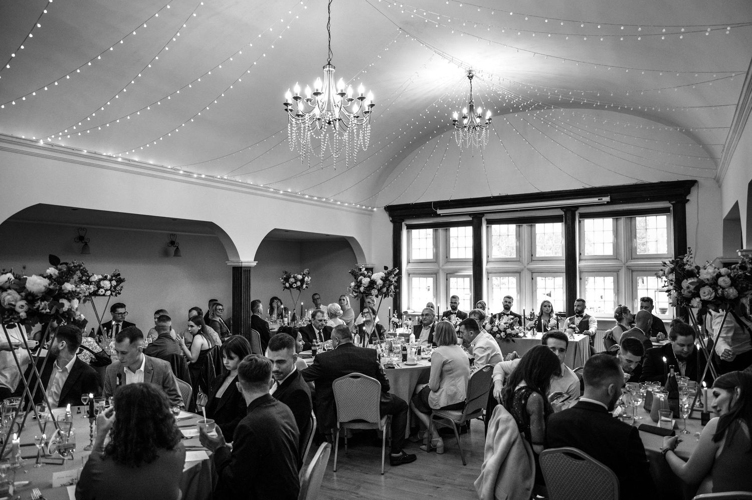 Czarno-białe ujęcie sali w restauracji Ołowianka w Gdańsku, goście weselni siedzący przy stołach, nad głowami świeci girlanda świetlna.