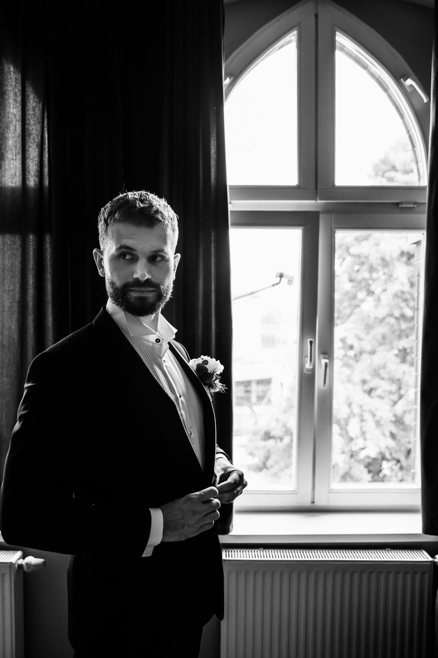 Portret Pana Młodego w eleganckim garniturze, uchwycony przy oknie w trakcie przygotowań — światło i kontrast w klasycznym stylu