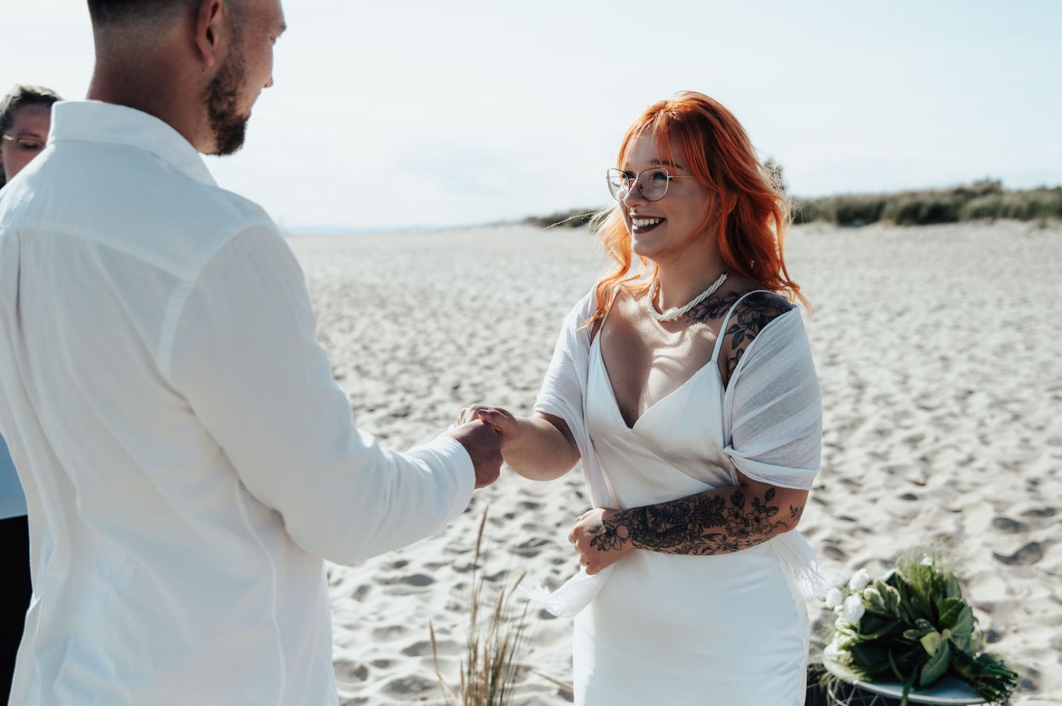 Romantyczny ślub na plaży – uśmiechnięta panna młoda w białej sukni i z rudymi włosami składa przysięgę ukochanemu na piasku w blasku słońca
