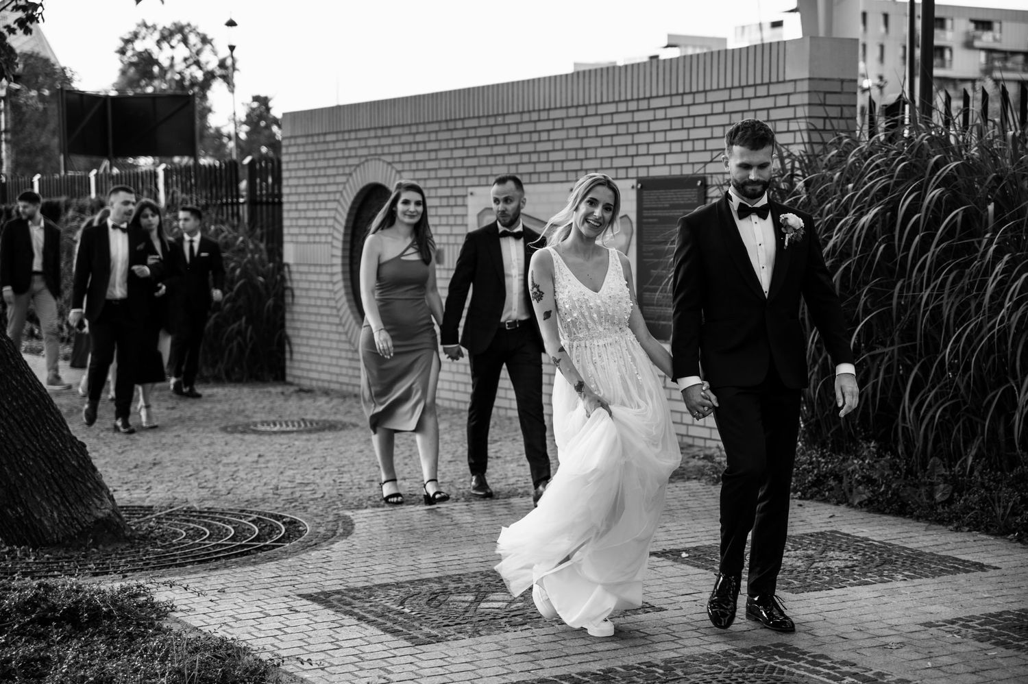 Para młoda idzie ulicą, prowadząc grupę gości w eleganckich stylizacjach – kadr w czerni i bieli.