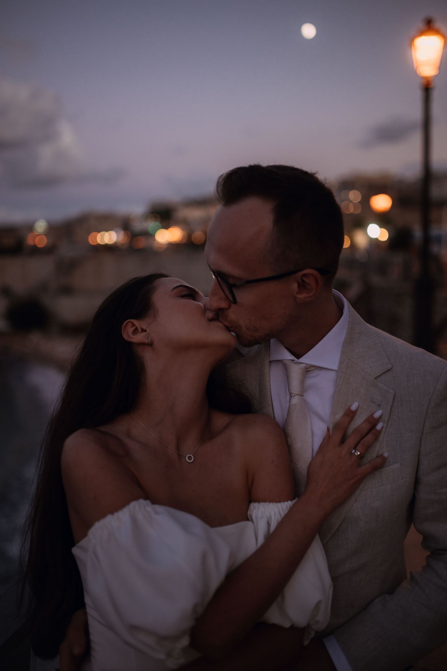 Romantyczny pocałunek pary młodej podczas sesji ślubnej w Castellammare del Golfo o zachodzie słońca, z księżycem i światłami w tle.