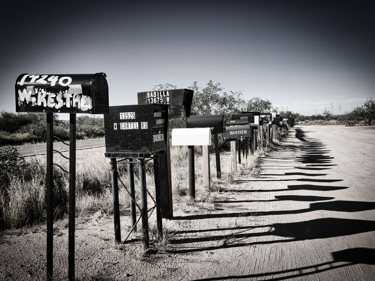 In the monochromatic scene, a row of vintage mailboxes stands solemnly by a quiet roadside. The desaturation emphasizes the textures