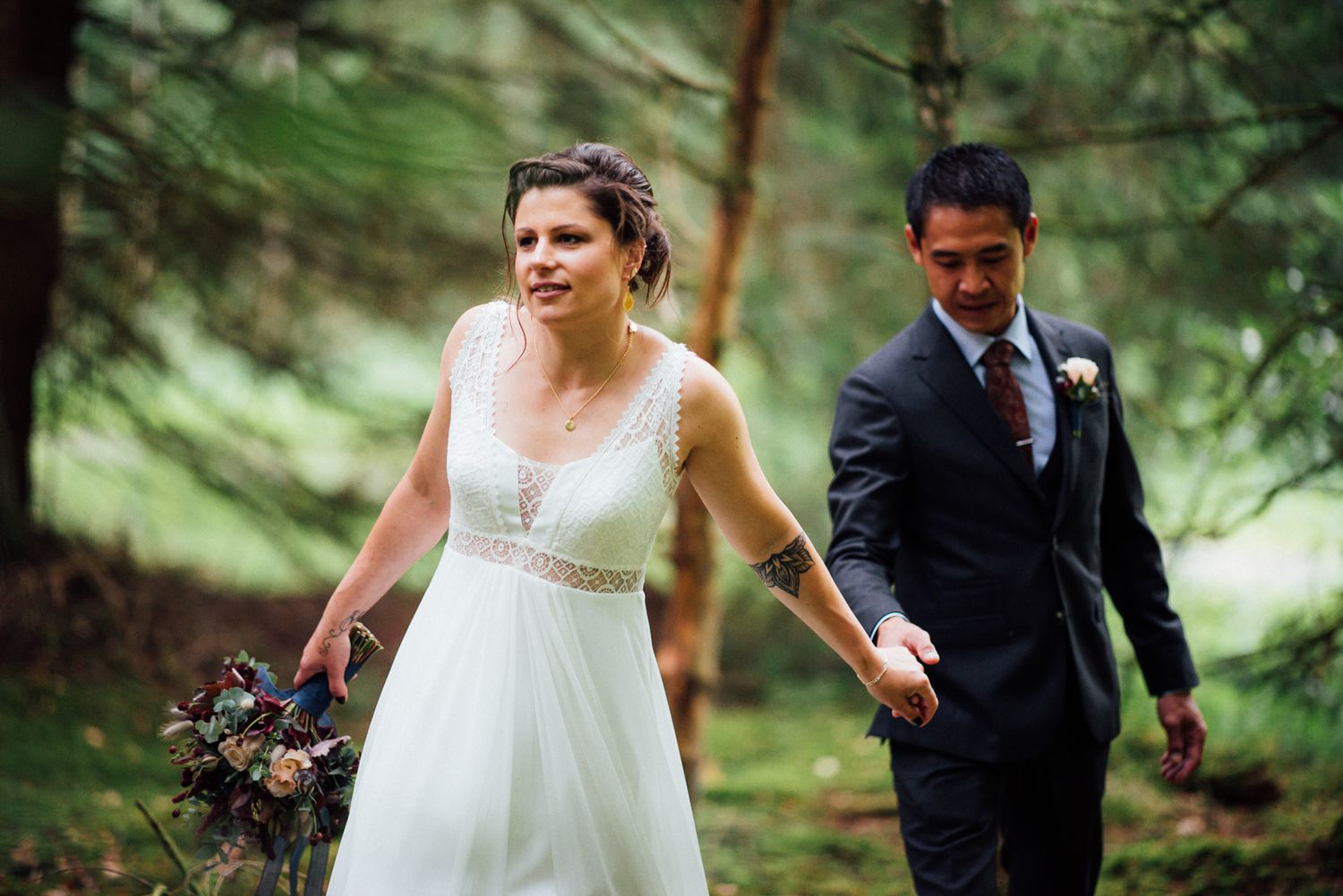 A bride in a white dress and groom in a dark suit walk through a lush forest setting during their wedding.