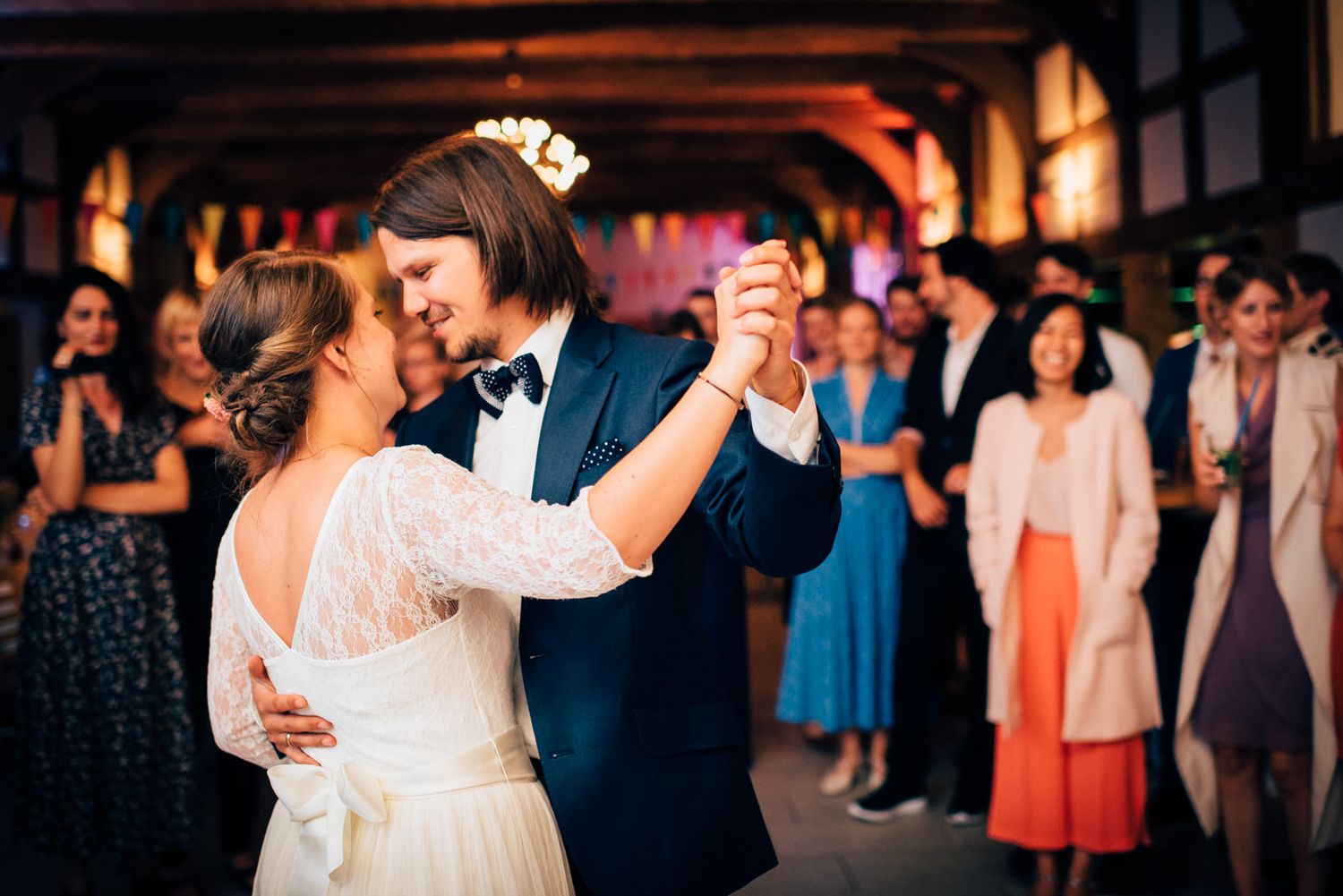 Dancing couple in formal attire surrounded by wedding guests in colorful dresses.