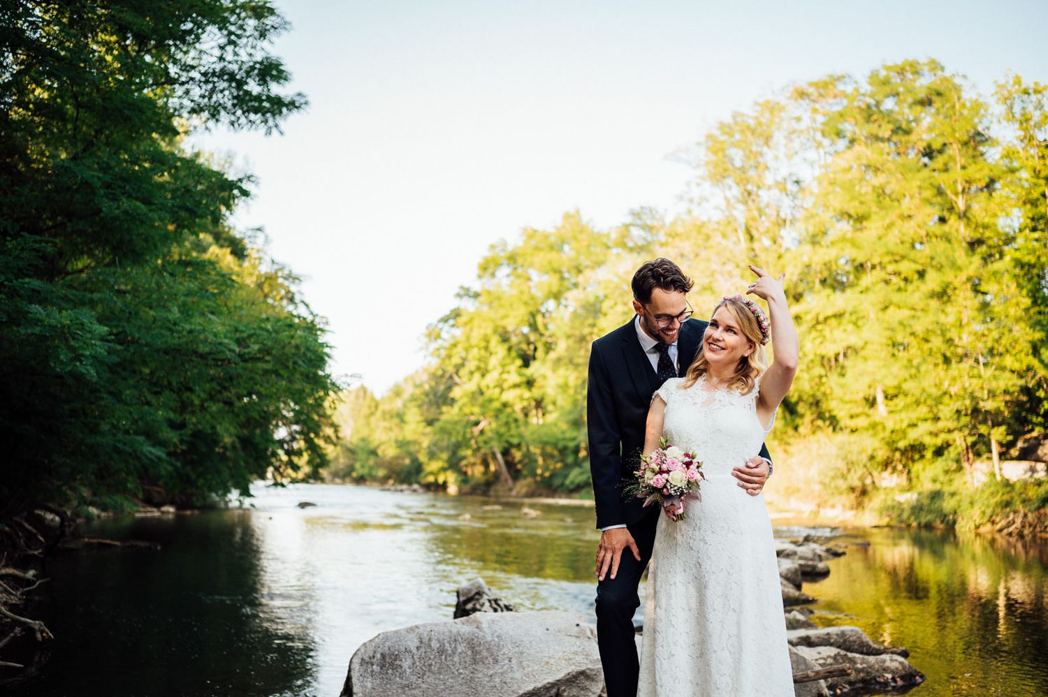 Wedding couple standing on rocks by a serene river with sunlit trees in the background.