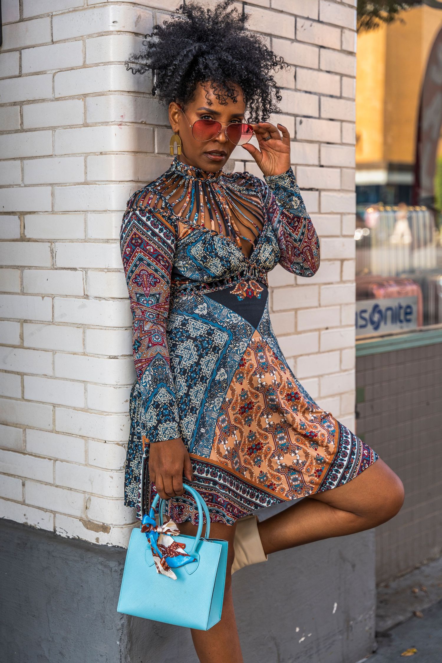 Fashion model poses against white brick wall wearing mixed print dress and holding turquoise handbag.