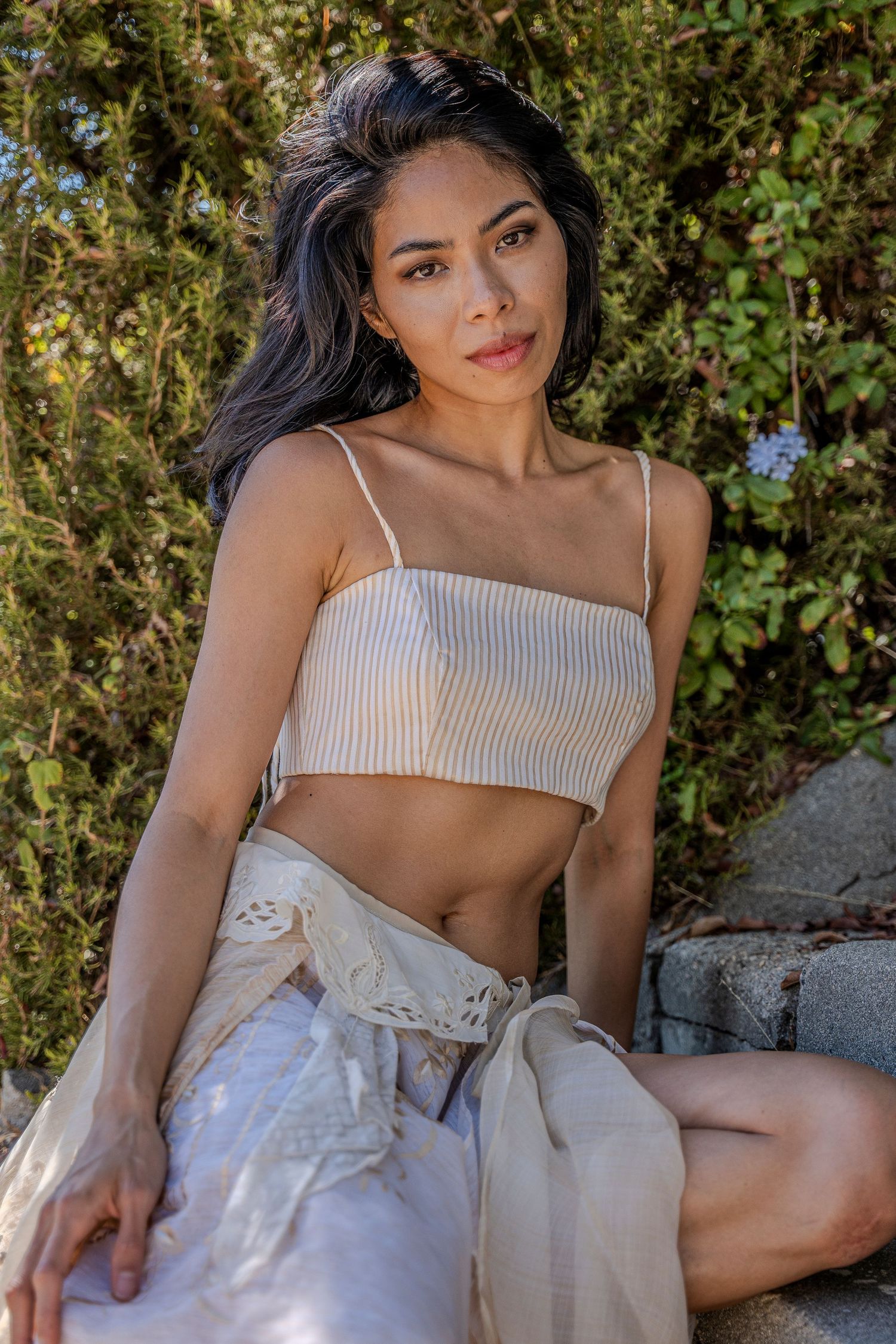 A woman in a white striped crop top and flowy skirt poses outdoors against a natural green backdrop.