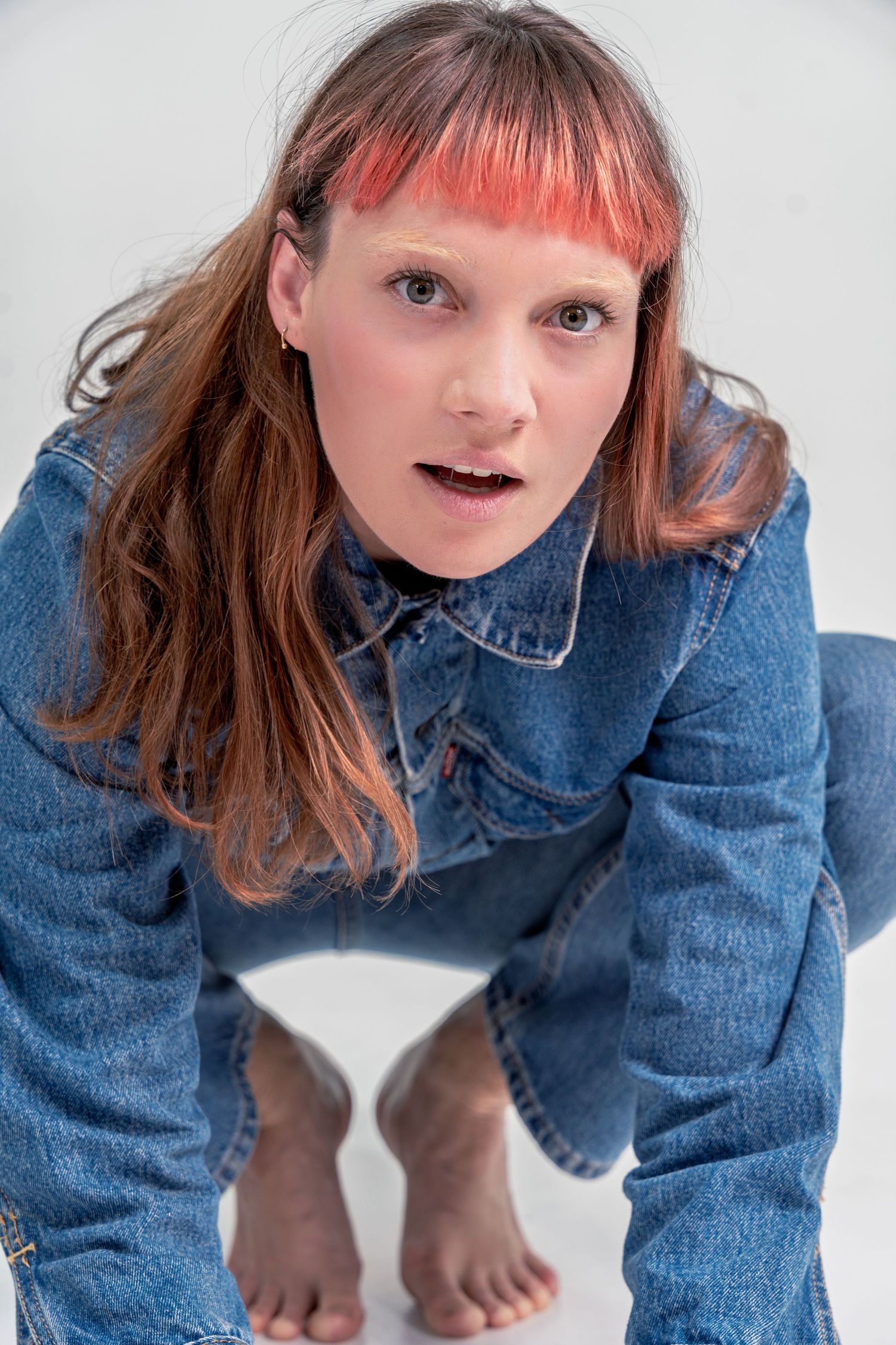 Person wearing denim jacket and red hair poses playfully in studio against white background.